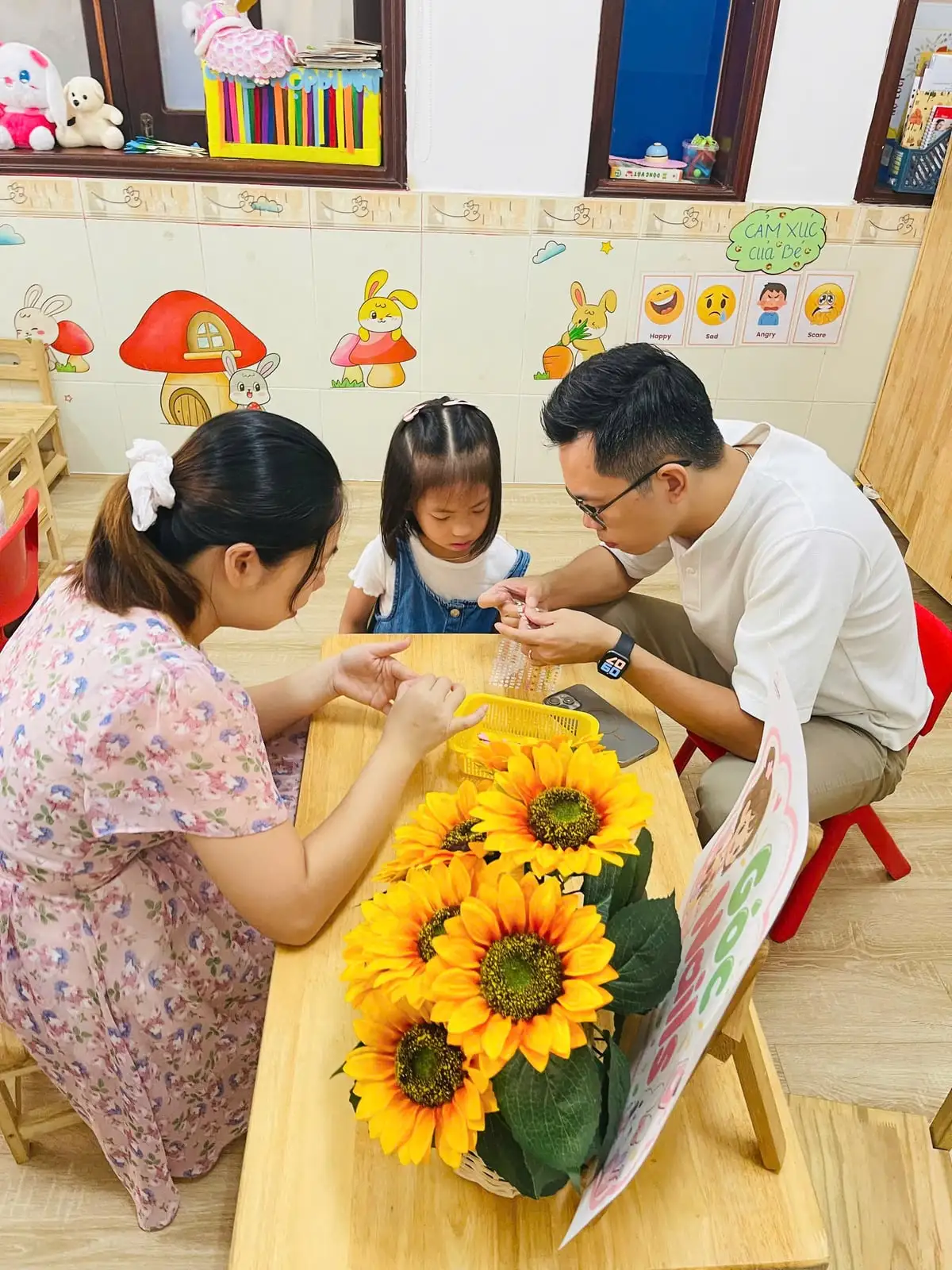 Ba mẹ cùng bé gái dán sticker trong lớp, phía sau là bảng "Cảm xúc của bé" với bốn khuôn mặt vui, buồn, giận, sợ