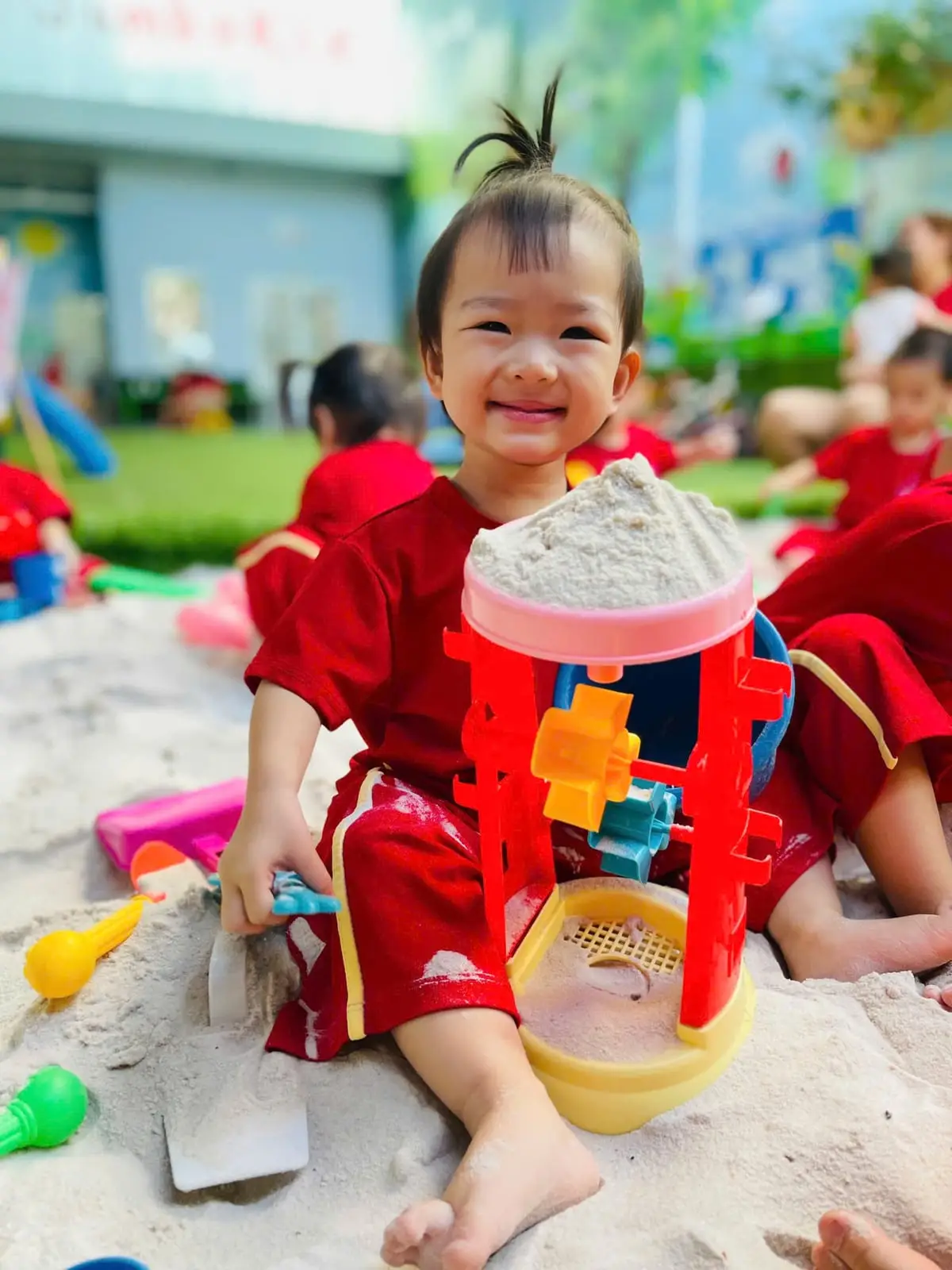 Bé gái tóc chỏm cười cầm khuôn cát ở khu chơi cát