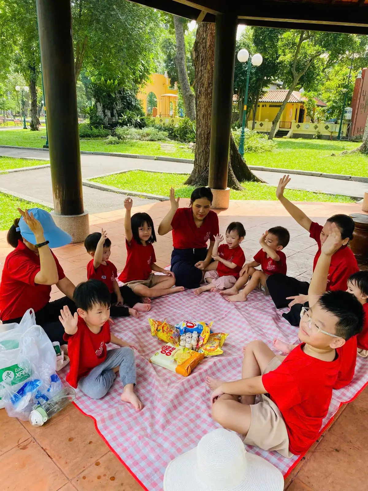 Các bé giơ tay hào hứng ngồi quanh tấm bạt trong buổi dã ngoại cùng cô