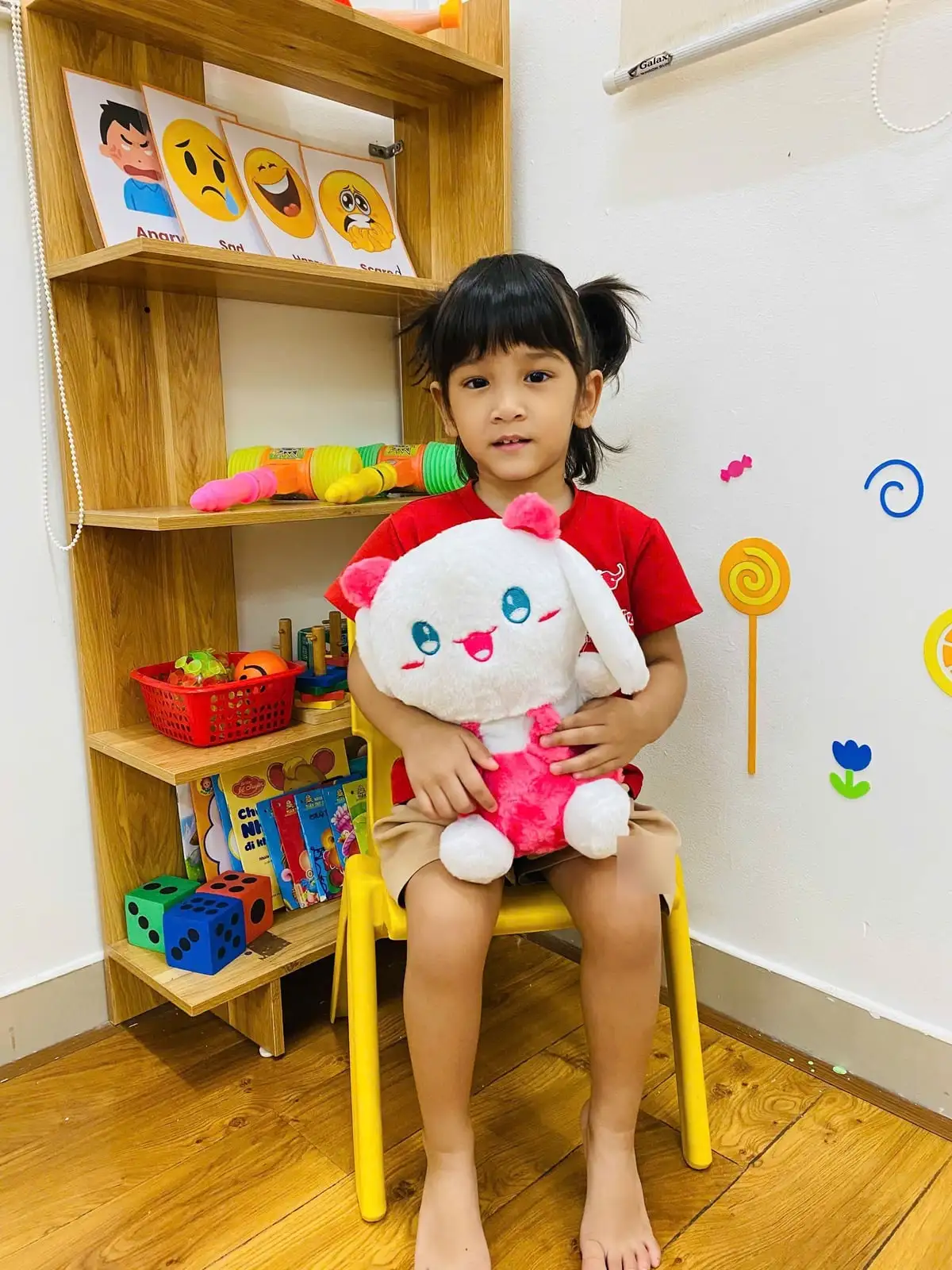 Bé gái mặc đồng phục đỏ ôm thỏ bông, ngồi ghế vàng ở góc lớp có thẻ cảm xúc