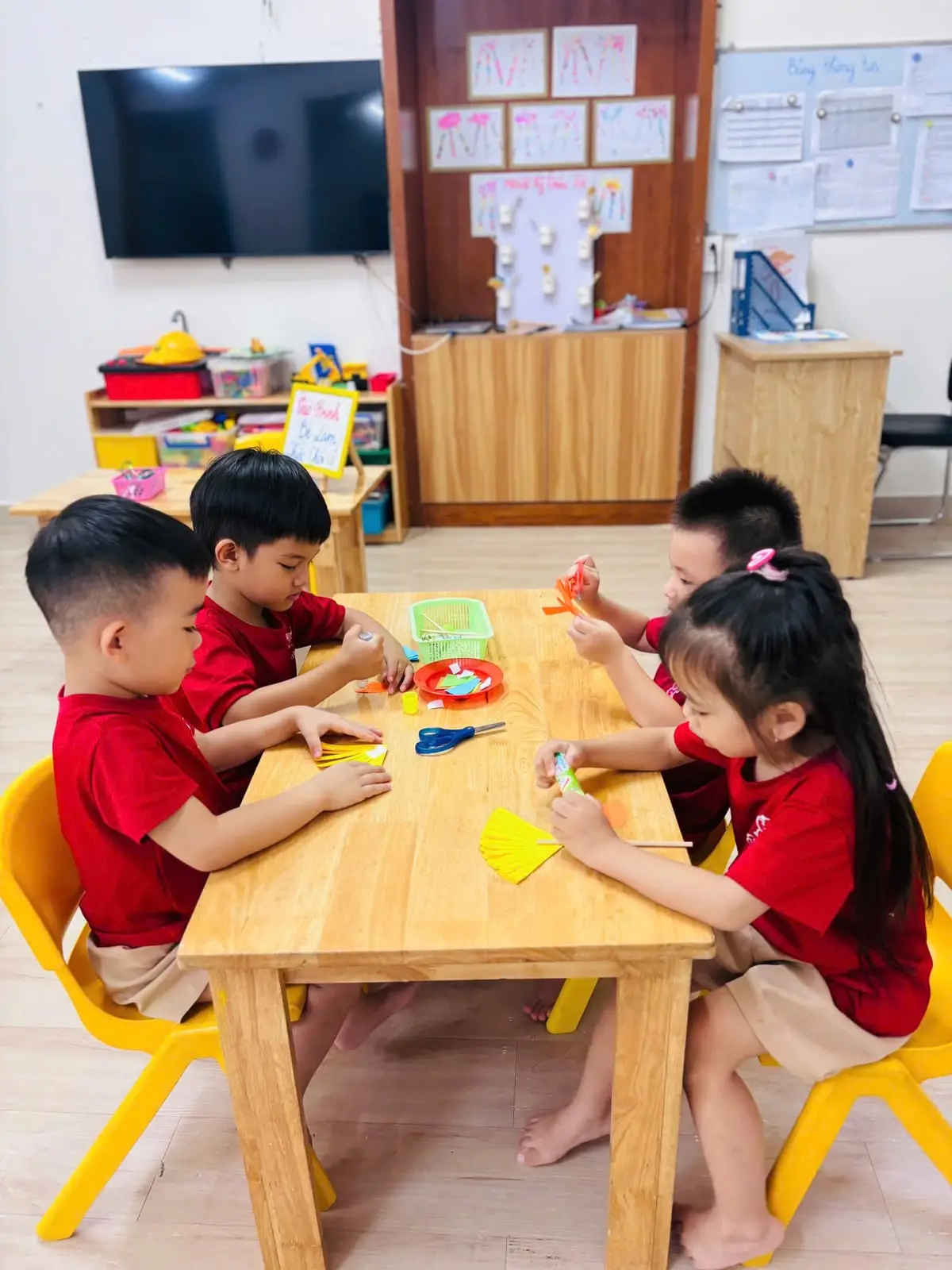 Bốn bé lớp Chồi mặc đồng phục đỏ ngồi quanh bàn gỗ, cùng cắt dán làm chổi giấy