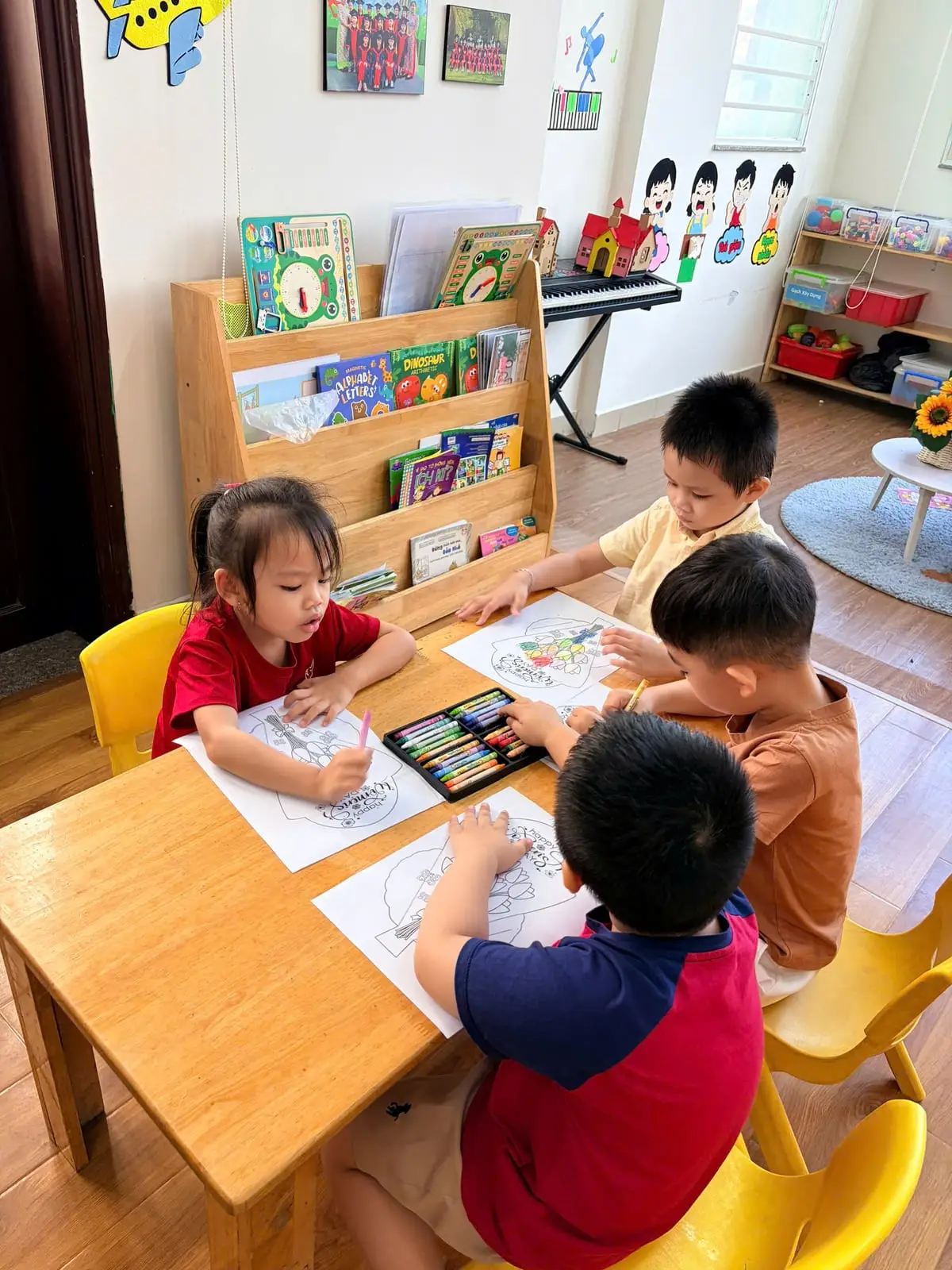 Bốn bé lớp Lá ngồi quanh bàn gỗ, cùng dùng chung hộp sáp màu để tô thiệp tặng mẹ