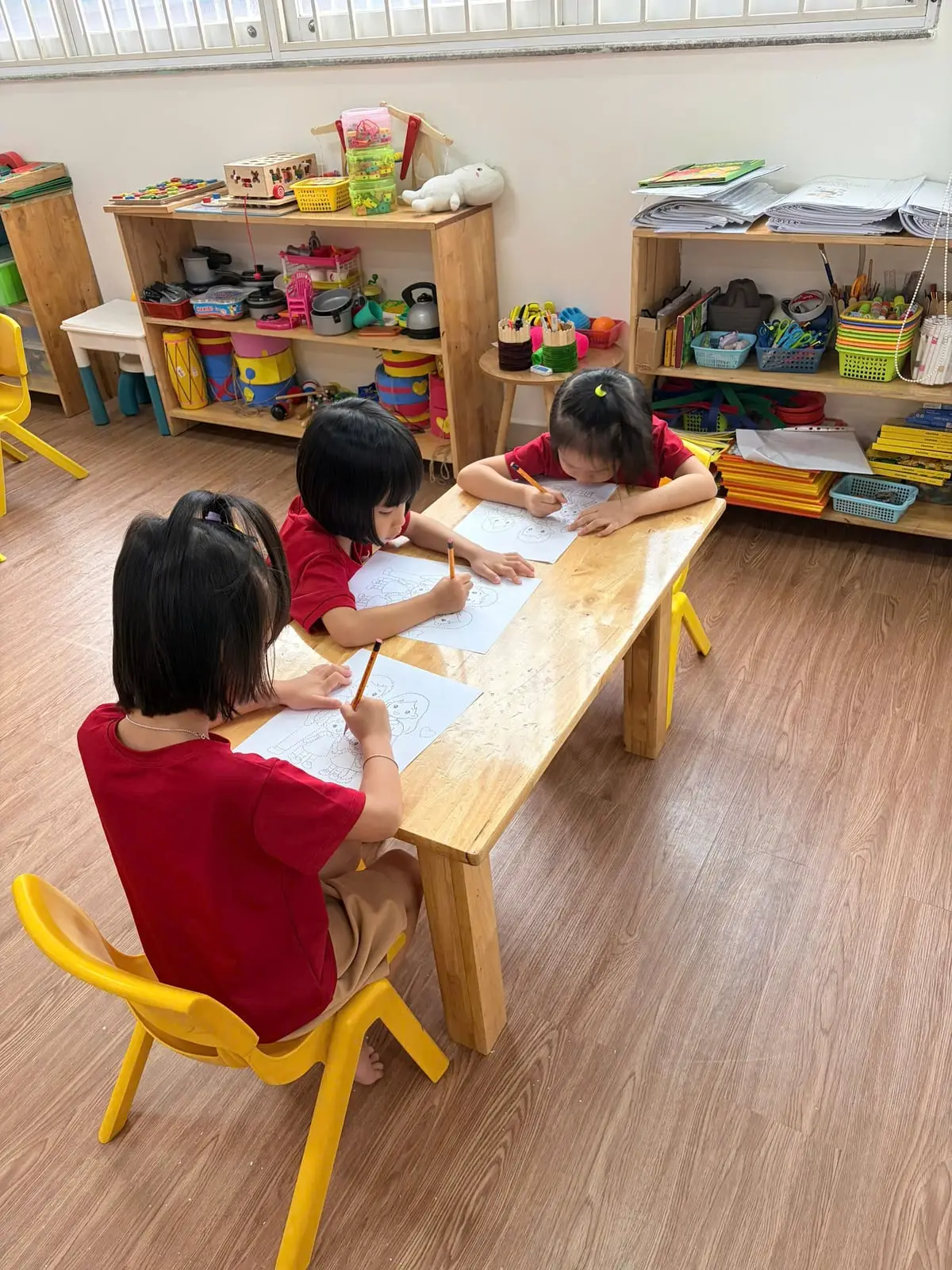 Ba bé gái lớp Lá ngồi bàn gỗ tô nét tranh trong lớp