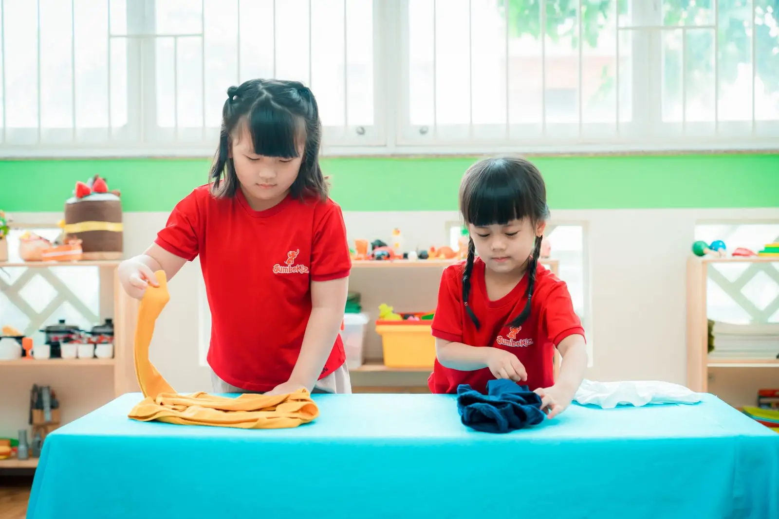 Hai bé gái tập gấp quần áo trên bàn, chuẩn bị cho trẻ vào lớp 1 bằng kỹ năng tự lập
