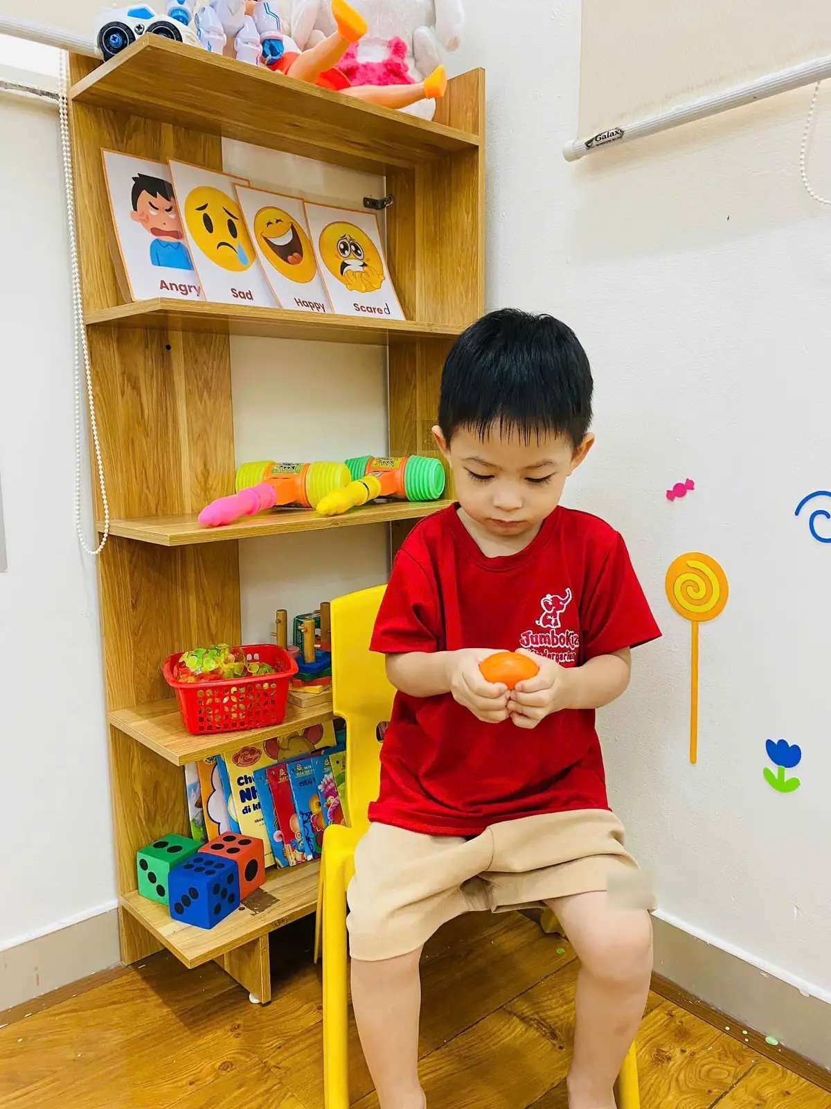 Bé trai ngồi ghế vàng, bóp quả bóng màu cam ở góc lớp có kệ thẻ cảm xúc