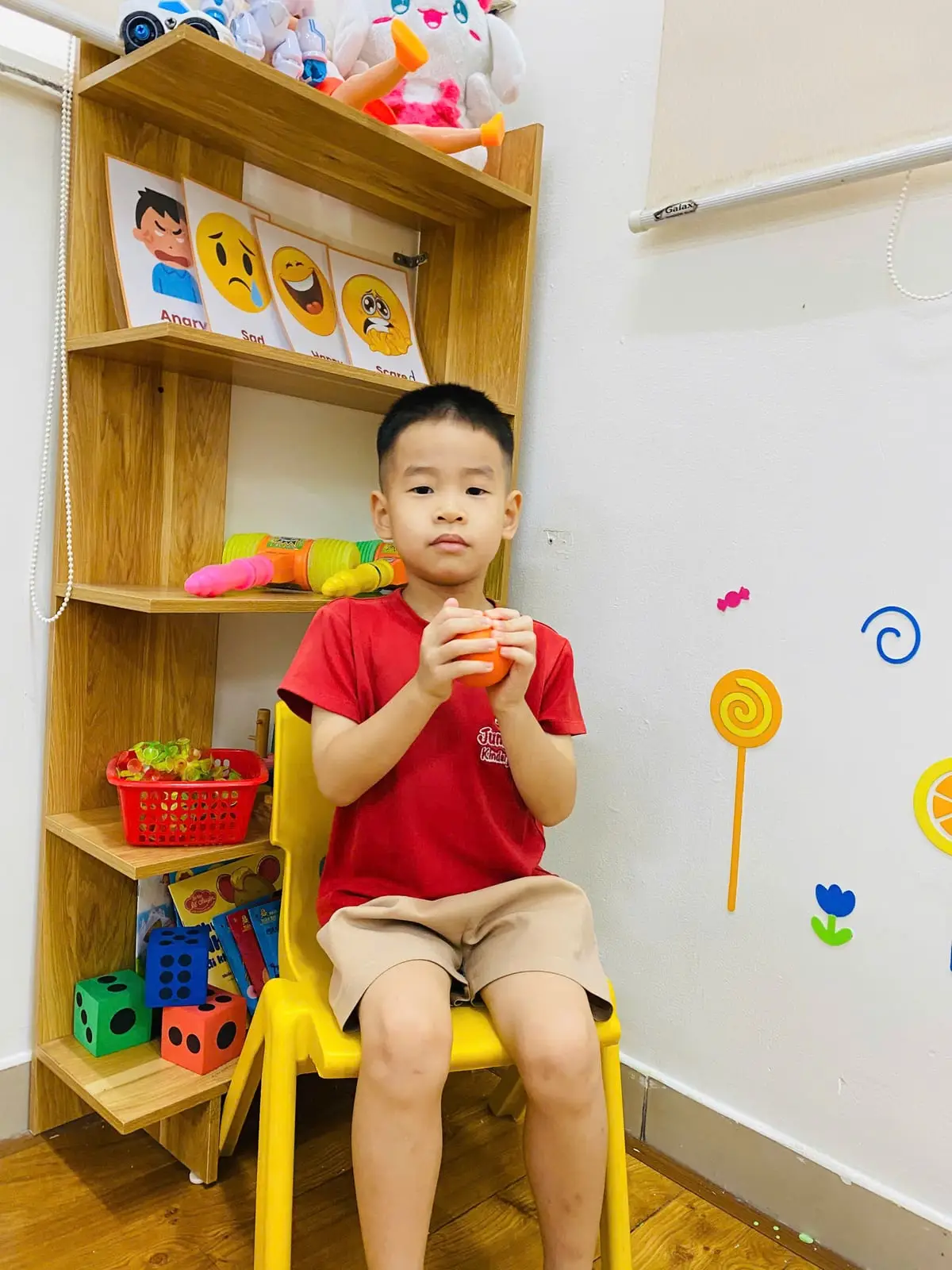 Bé trai mặc đồng phục đỏ cầm quả bóng mềm, ngồi ghế vàng ở góc lớp có thẻ cảm xúc