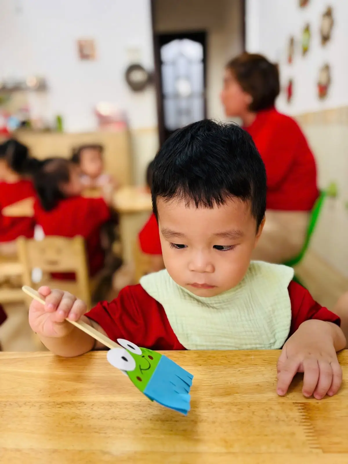 Bé trai đeo yếm ngồi ở bàn, cầm chiếc chổi ếch giấy thủ công