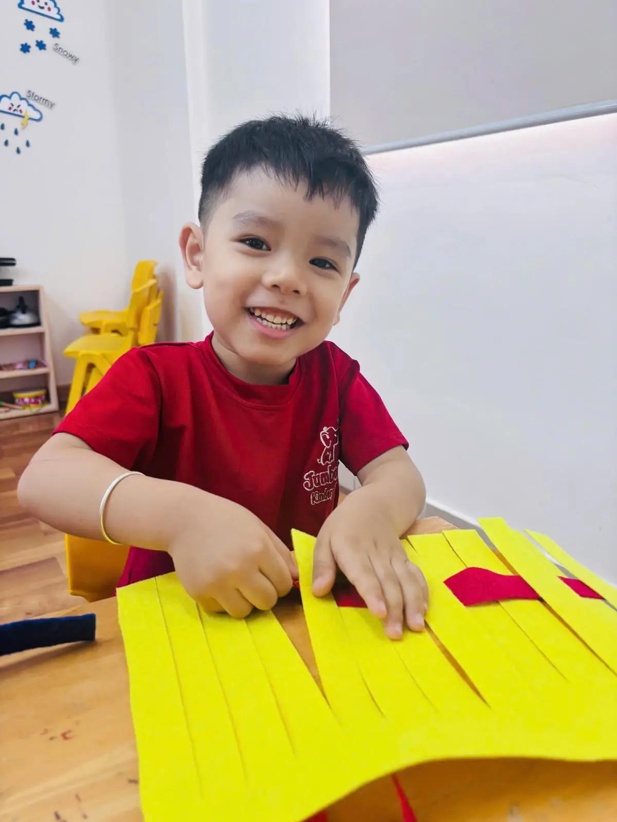Bé trai mặc đồng phục đỏ cười tươi bên tấm đan nỉ vàng trong giờ thủ công