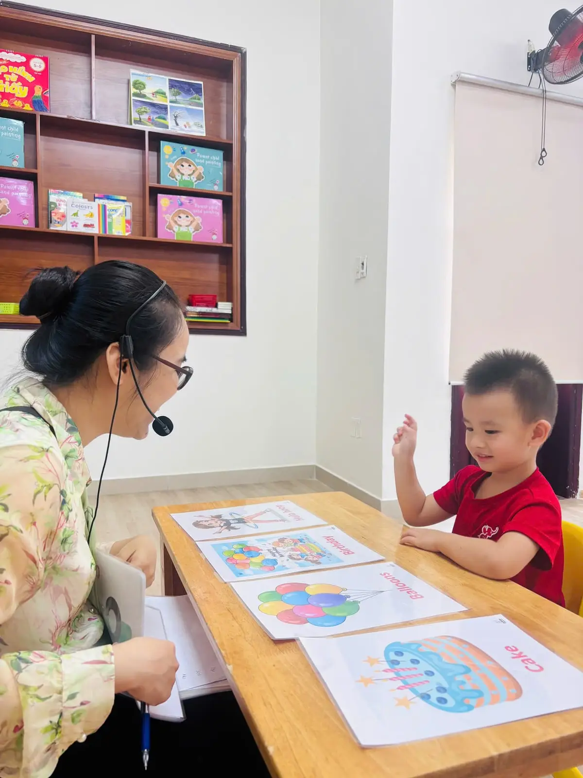 Bé trai cười, giơ tay trả lời cô bên bàn thẻ tranh
