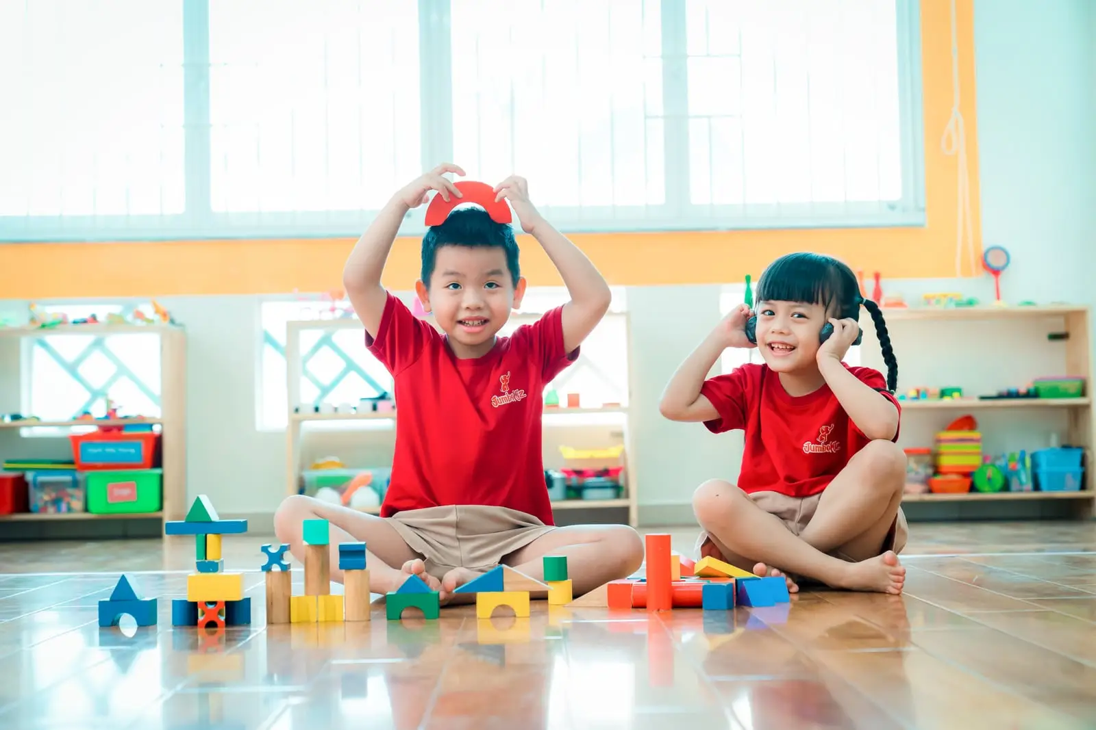 Hai bé ngồi trên sàn lớp xếp khối gỗ và cười đùa