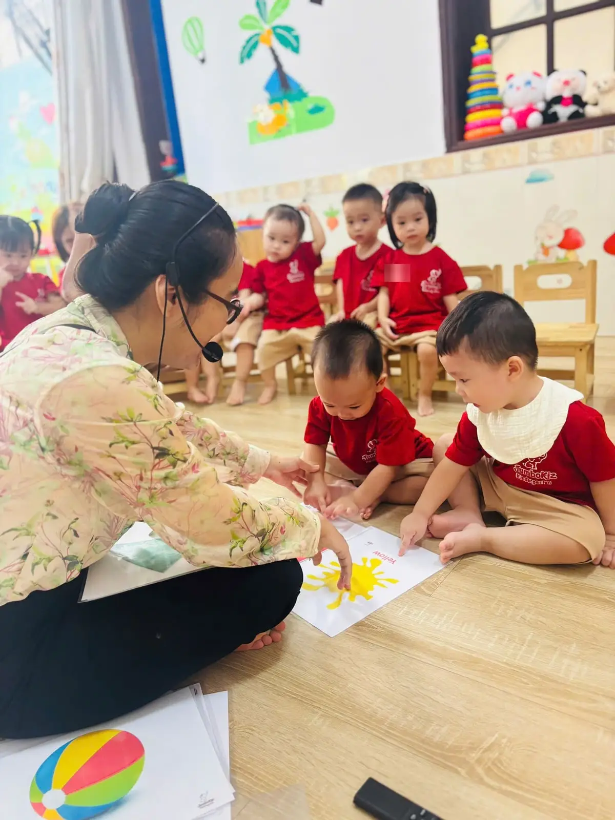 Cô ngồi trên sàn chỉ tranh màu vàng cho các bé lớp Nhà trẻ