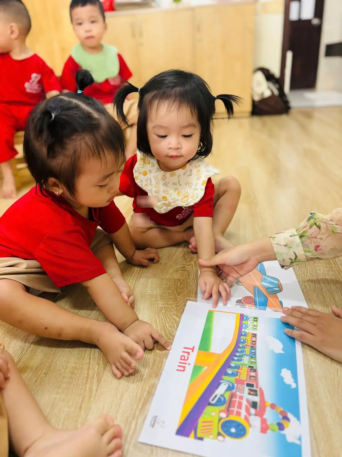 Cô cùng hai bé lớp Nhà trẻ chỉ tranh tàu hỏa trên sàn lớp