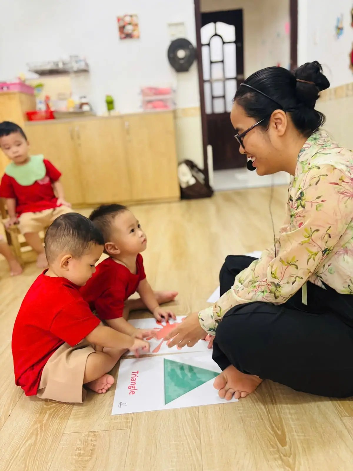 Cô ngồi cạnh, cười với hai bé lớp Nhà trẻ đang chạm tay vào thẻ hình