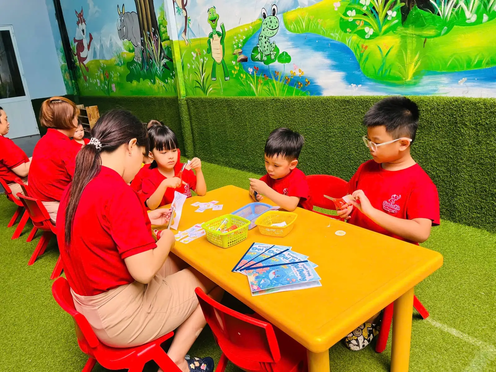 Cô ngồi cùng bàn hướng dẫn các bé cắt dán