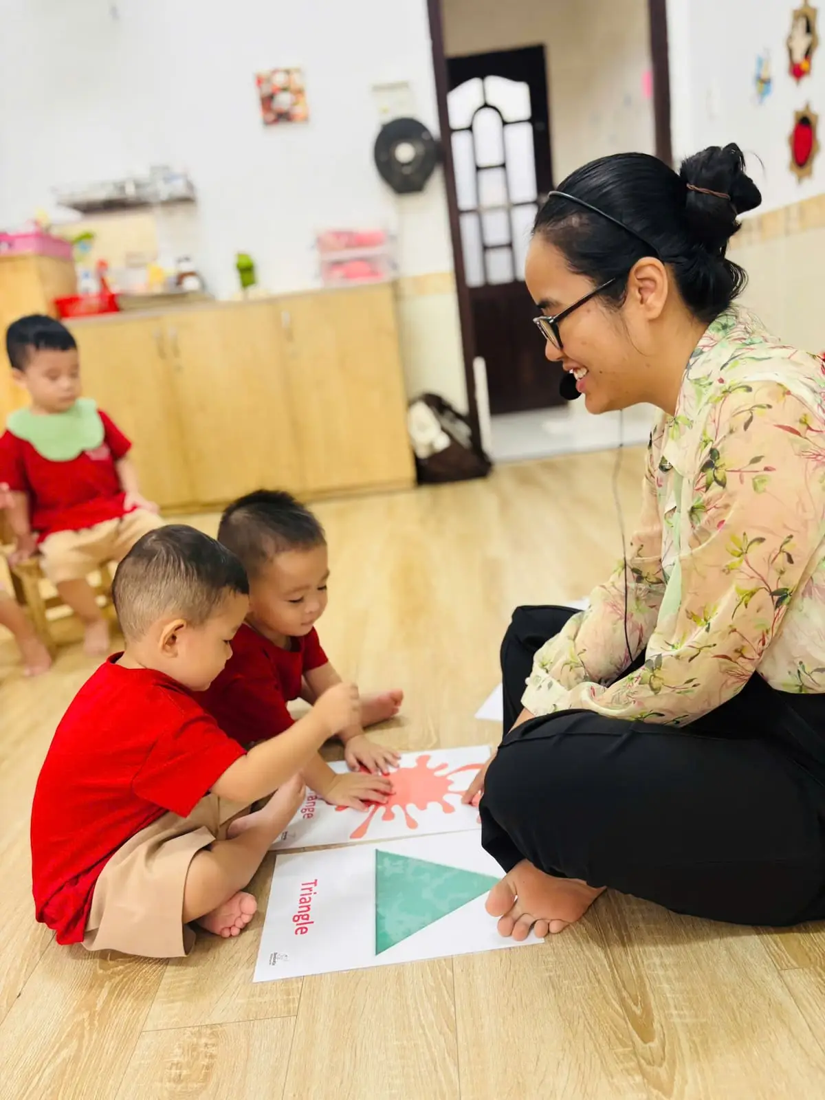 Cô ngồi ngang tầm hai bé ở lớp nhà trẻ, cùng con xem thẻ hình