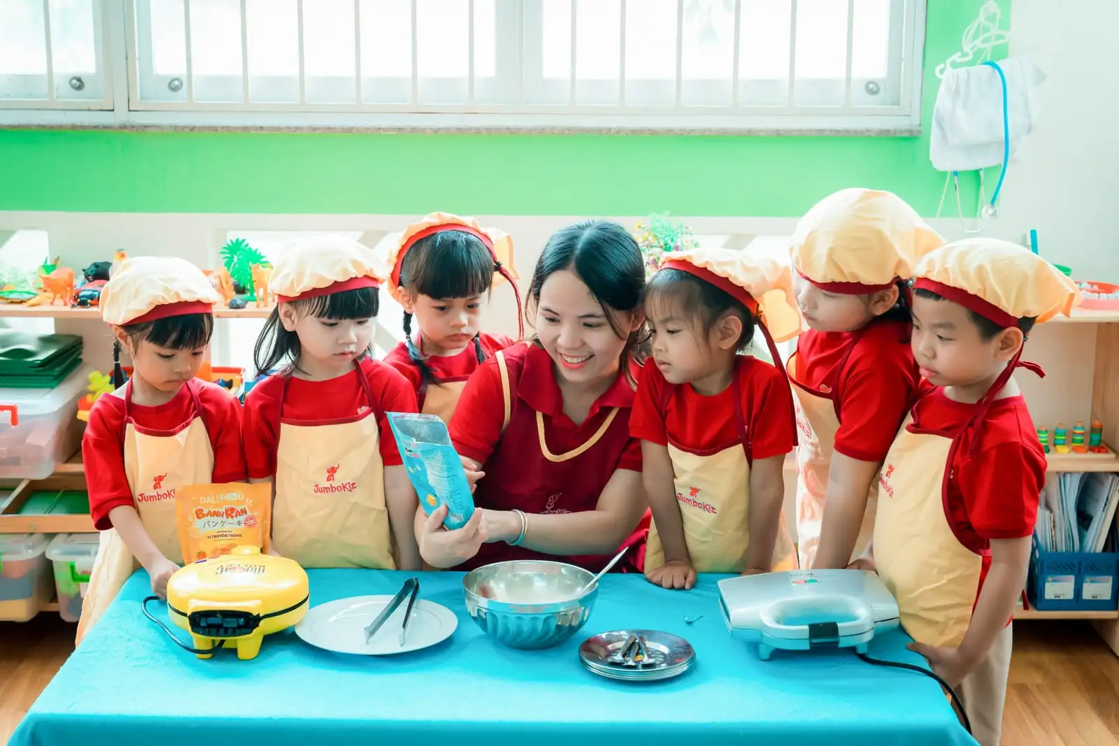 Cô giáo JumboKiz cùng sáu bé đội mũ đầu bếp chuẩn bị làm bánh trong lớp