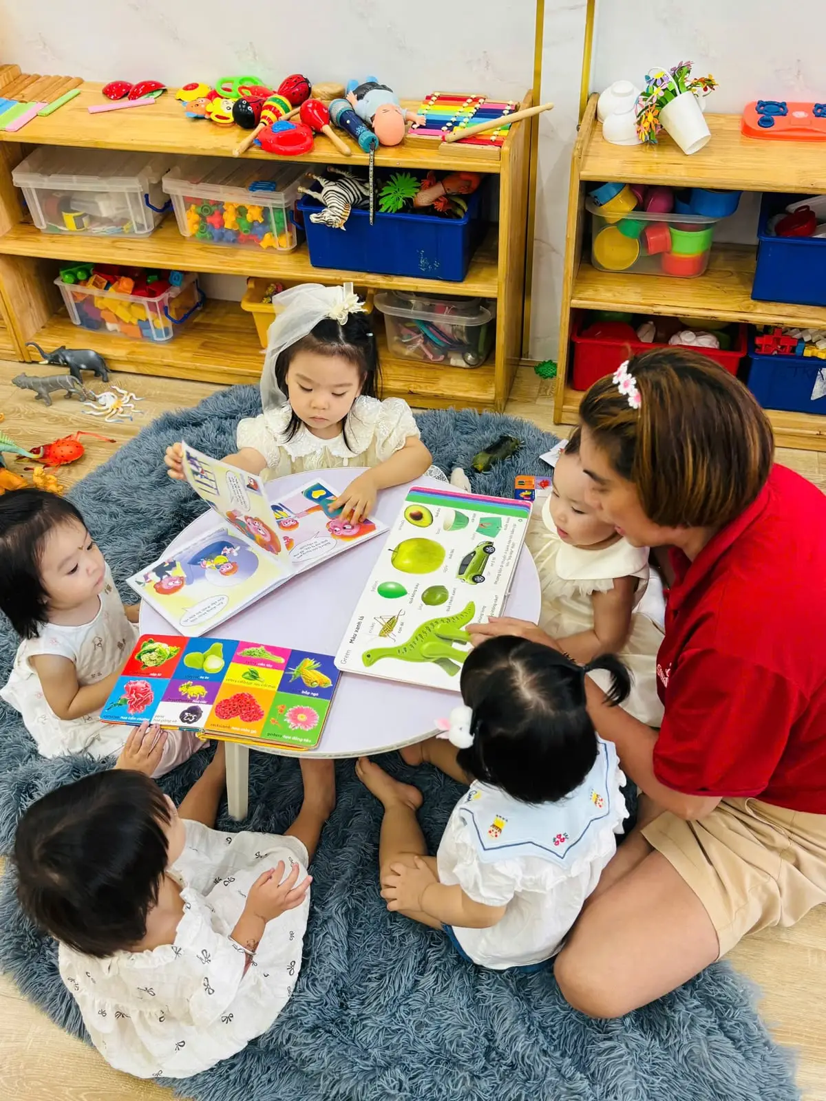 Cô và nhóm bé nhà trẻ ngồi quanh bàn tròn cùng xem sách tranh