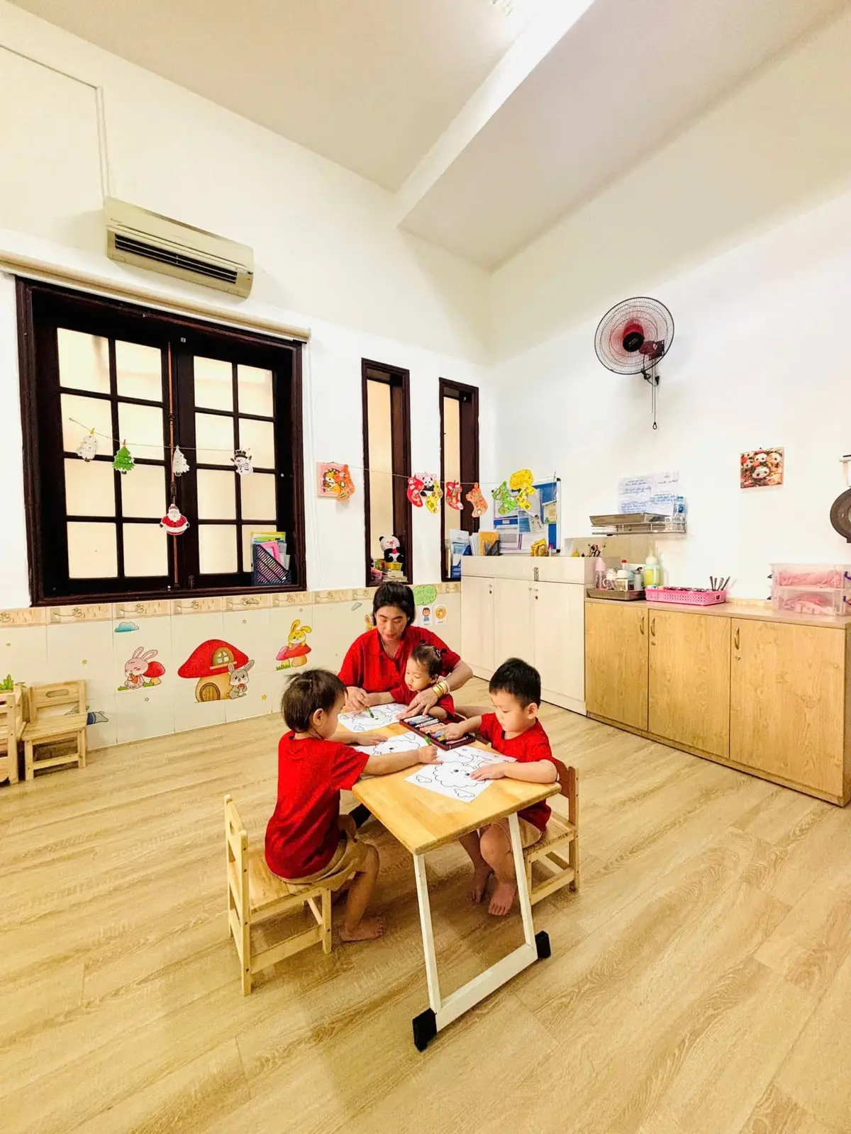 Cô ngồi cùng ba bé tô màu ở bàn gỗ trong phòng học rộng