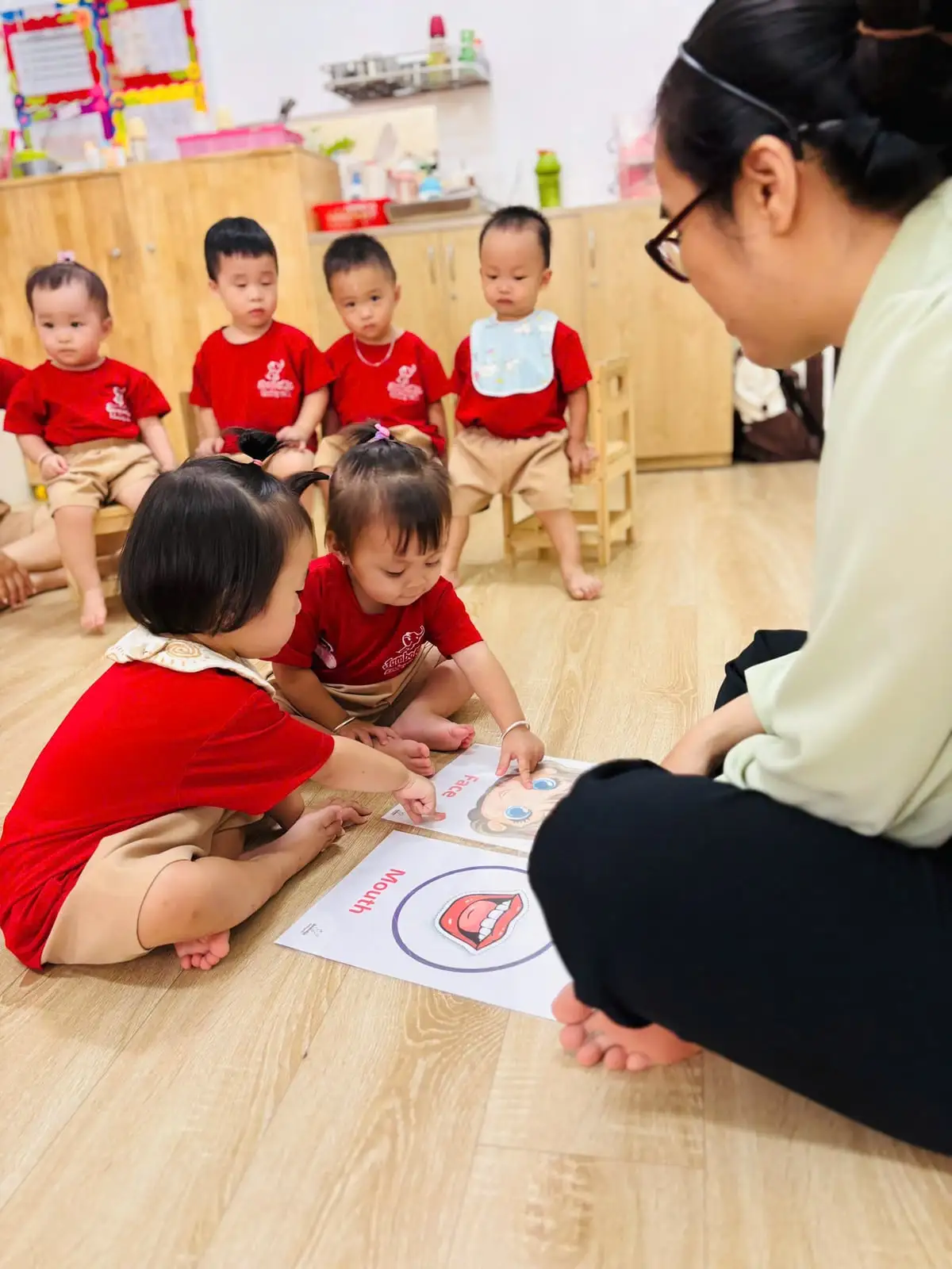 Cô và các bé lớp Nhà trẻ ngồi sàn cùng xem thẻ hình khuôn mặt, cái miệng
