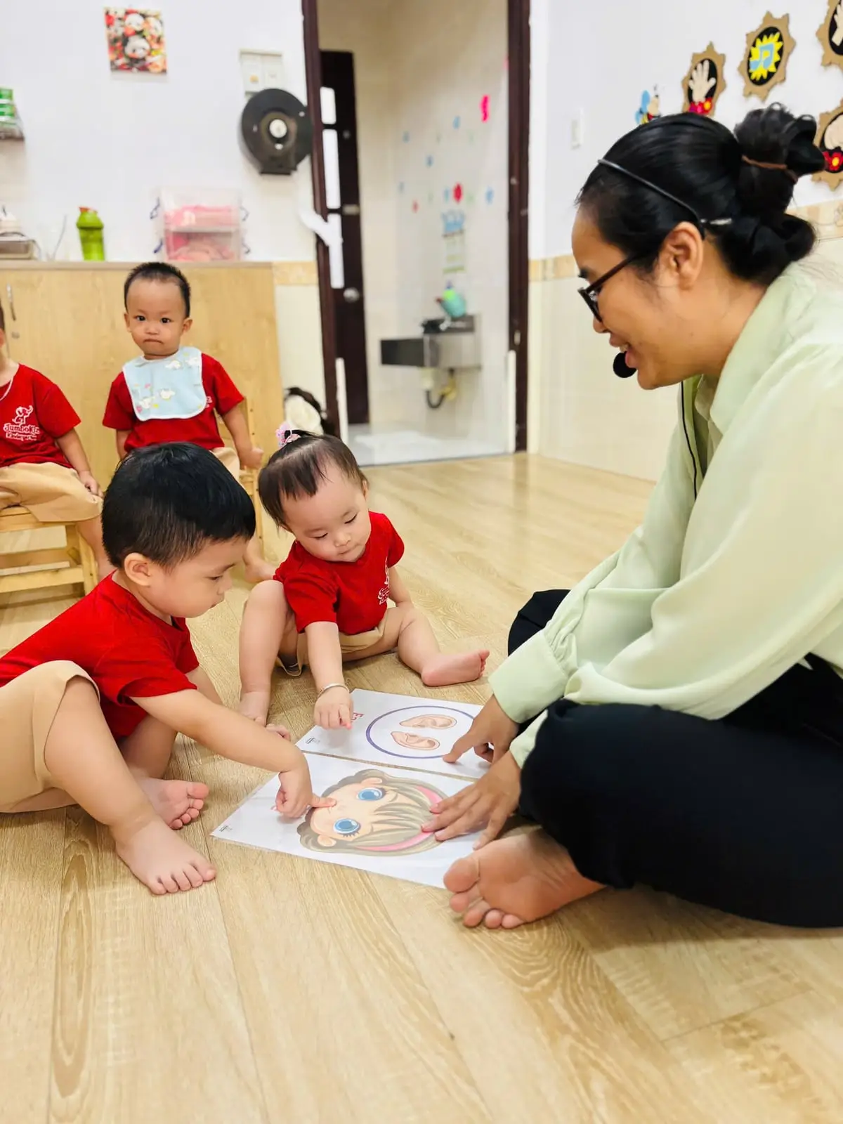 Cô ngồi ngang tầm hai bé ở lớp nhà trẻ, cùng con xem thẻ hình khuôn mặt