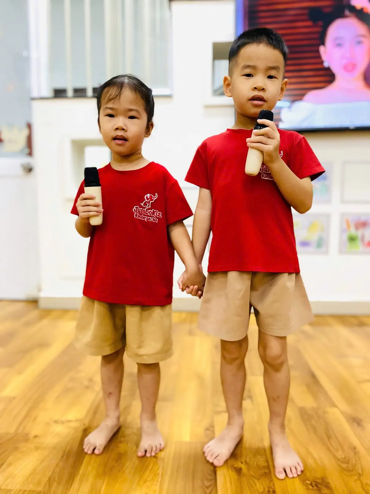Hai bé mặc đồng phục đỏ nắm tay nhau, cầm micro tập hát trong lớp