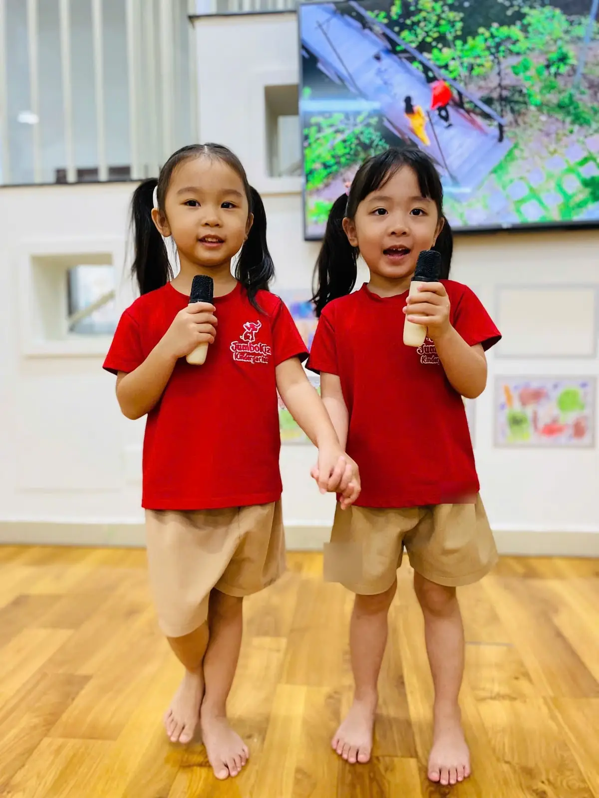 Hai bé gái mặc áo đỏ đồng phục nắm tay nhau, mỗi bé cầm một chiếc micro và hát