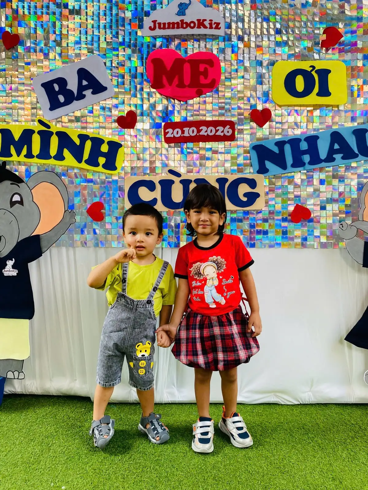 Hai bé nắm tay nhau chụp ảnh trước phông chữ Ba Mẹ Ơi