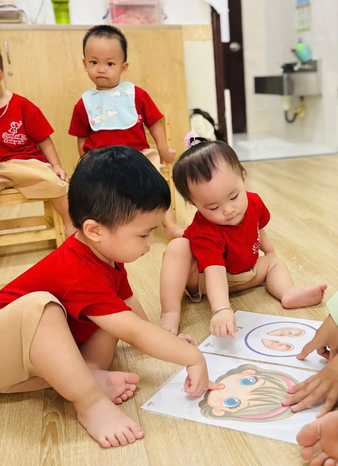 Hai bé lớp Nhà trẻ ngồi trên sàn lớp, chỉ vào thẻ hình khuôn mặt