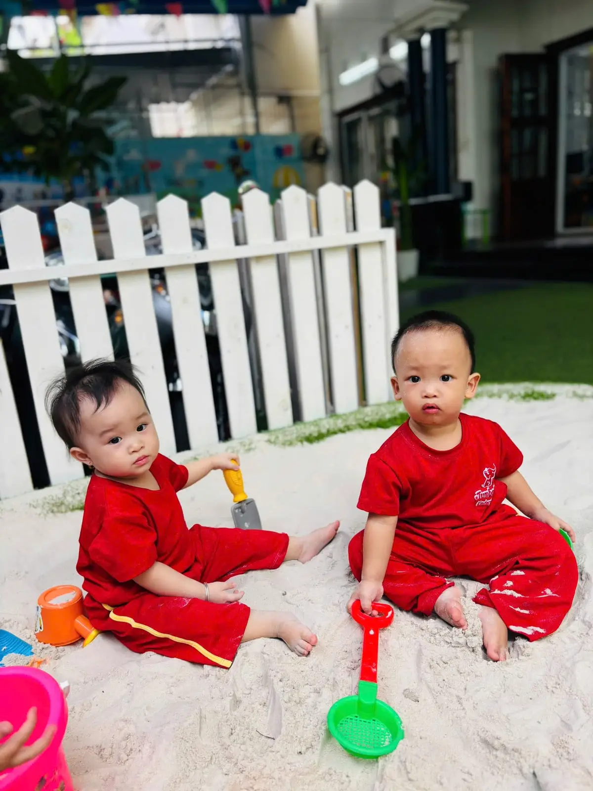 Hai bé lớp Nhà trẻ ngồi chơi cát cạnh nhau bên hàng rào trắng