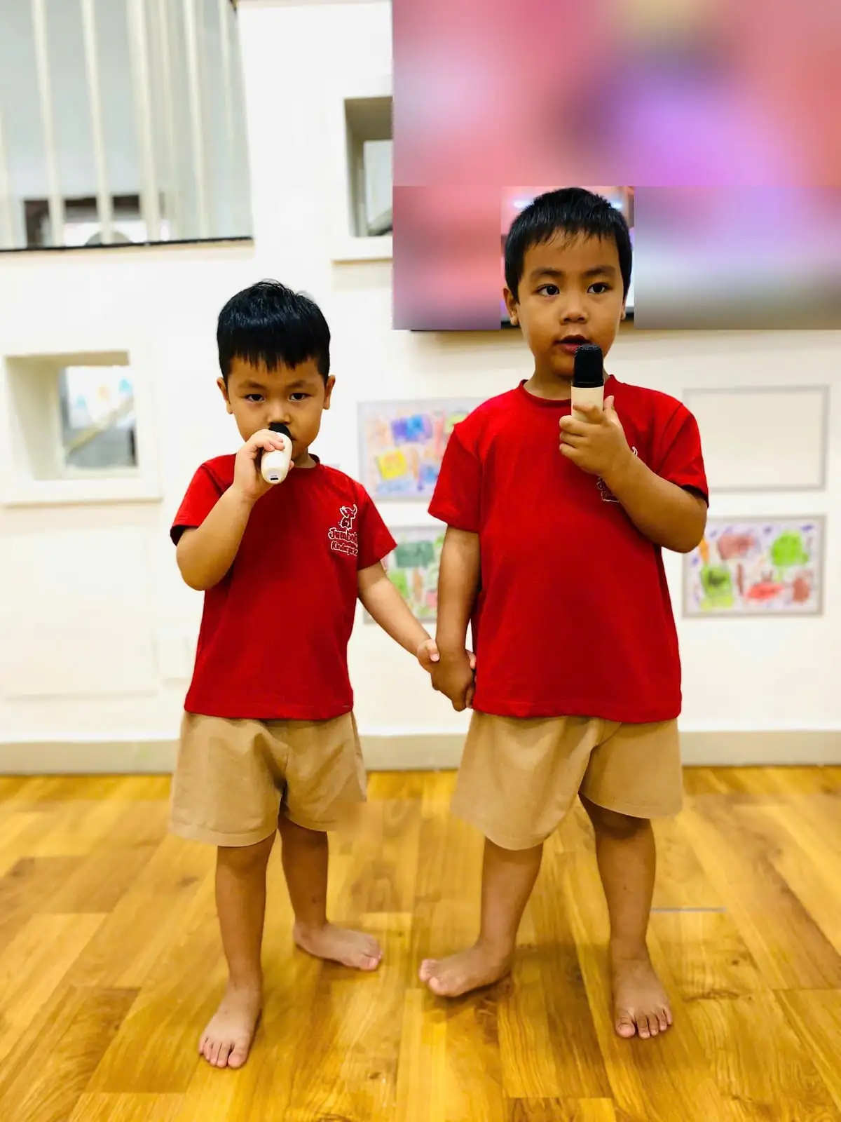 Hai bé trai nắm tay nhau, mỗi bạn cầm một micro tập hát trong lớp