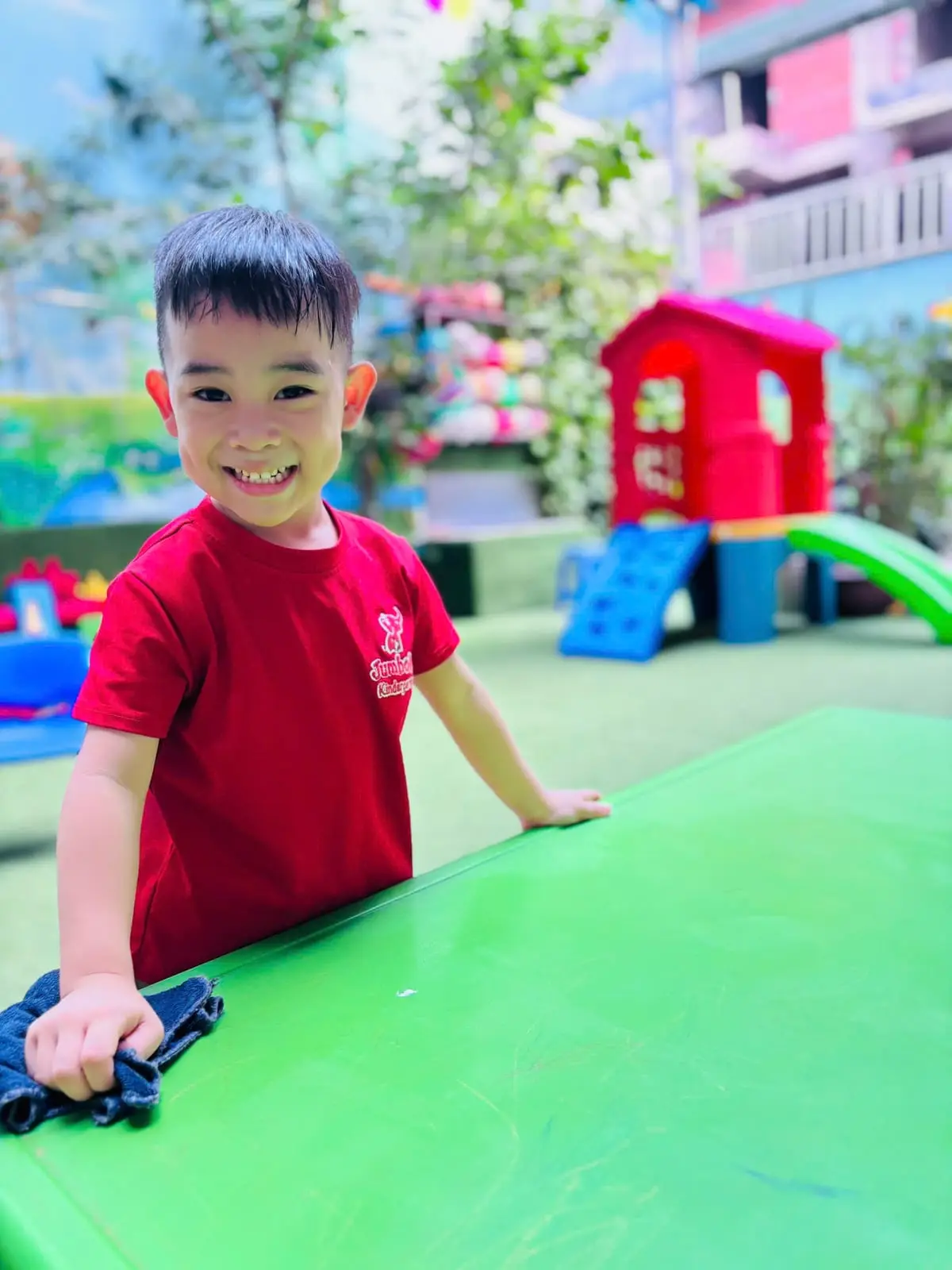 Bé trai lớp Chồi K2 cười khi lau bàn ở sân chơi