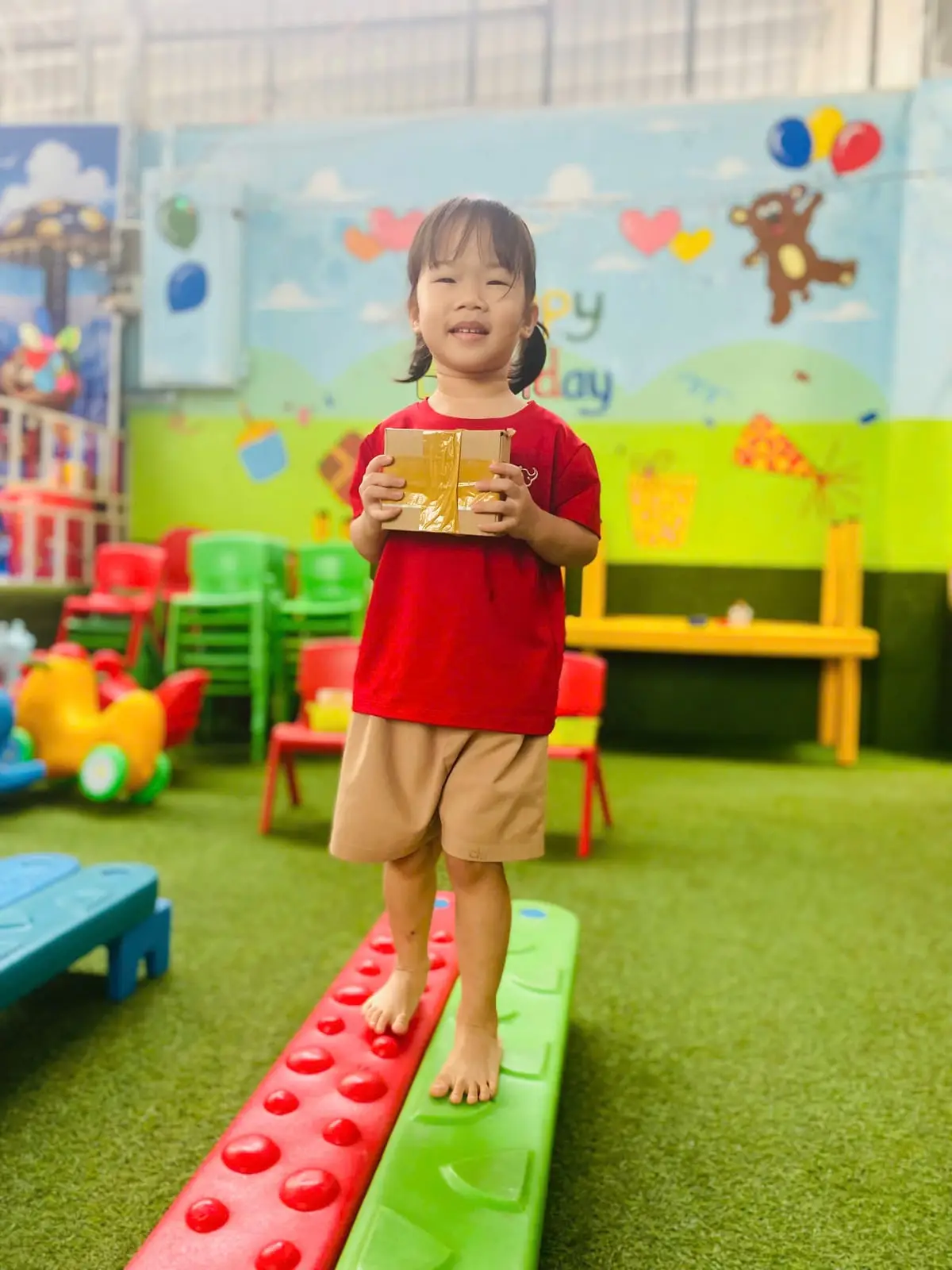 Bé gái lớp Mầm K1 cười, ôm hộp đi trên cầu thăng bằng đỏ xanh