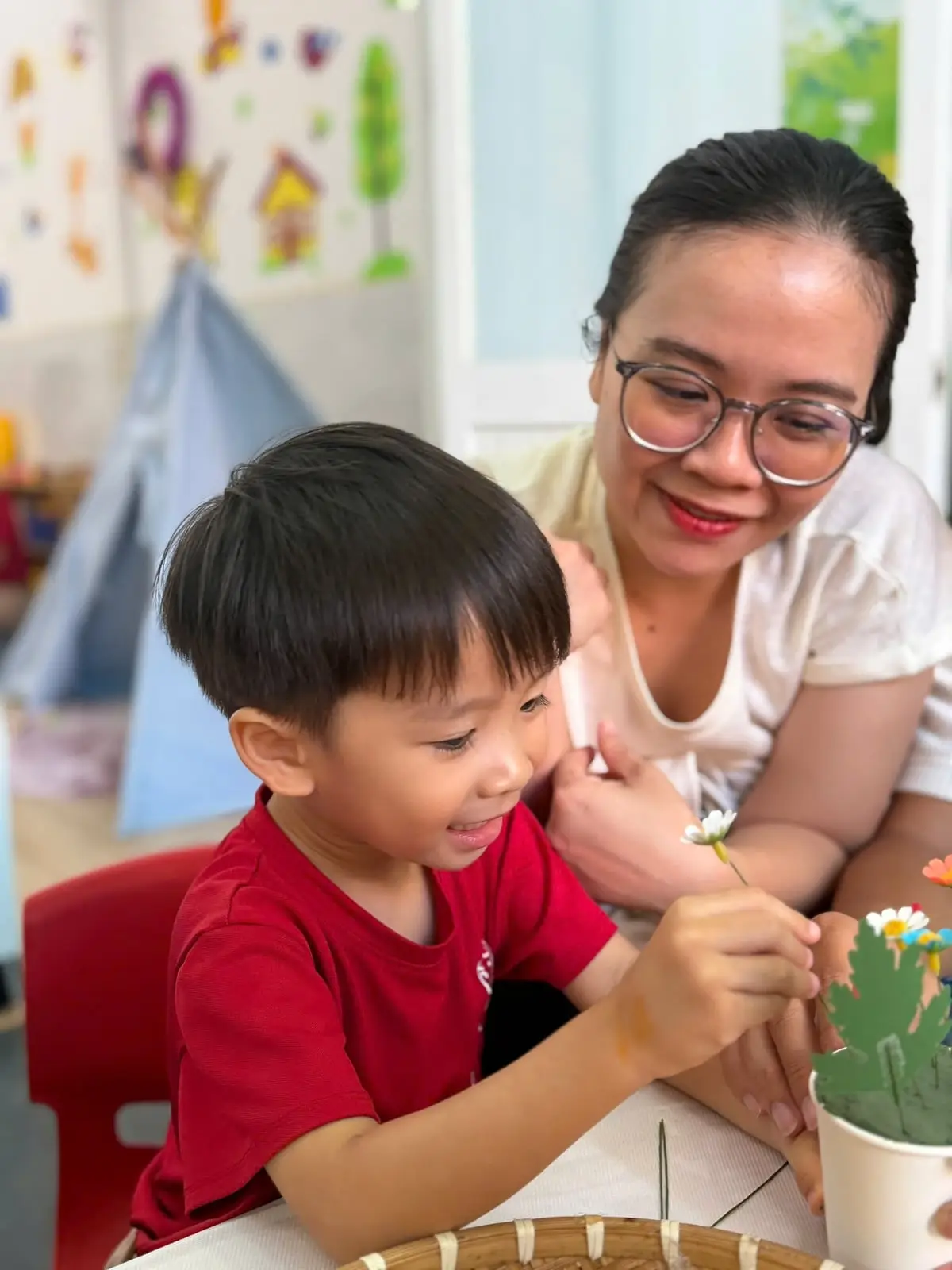 Mẹ mỉm cười ngồi cạnh, nhìn con trai tự cắm hoa vào cốc