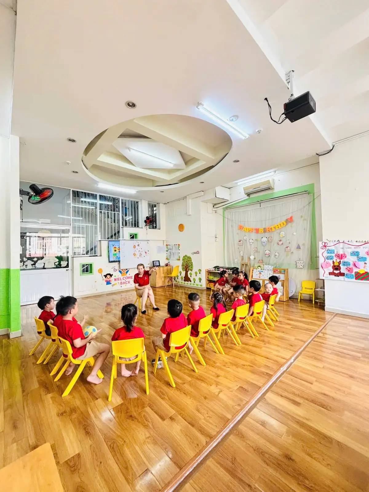 Các bé ngồi ghế vàng thành vòng tròn nghe cô trong lớp JumboKiz