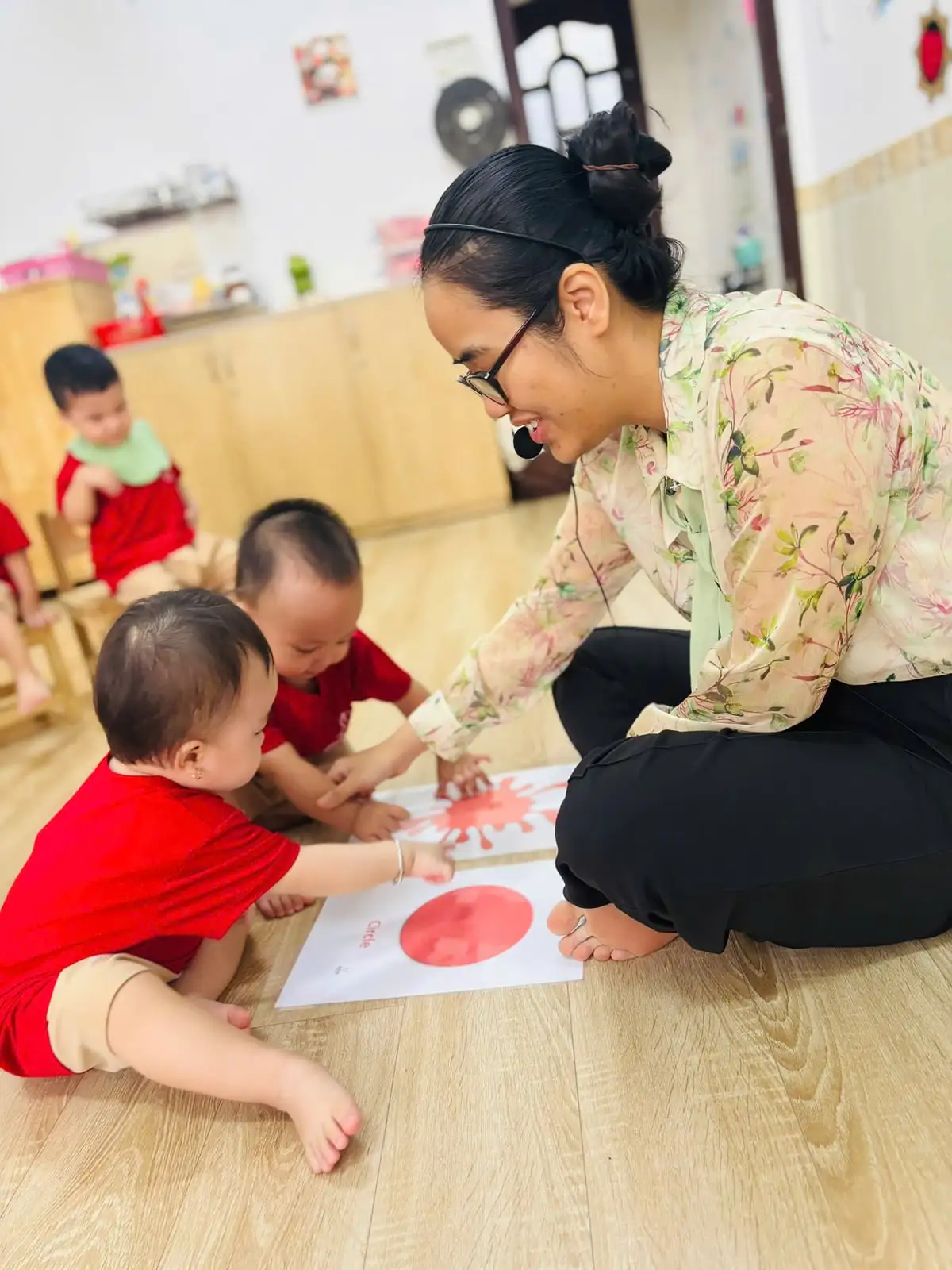 Cô ngồi trên sàn cùng hai bé lớp Nhà trẻ học hình tròn màu đỏ