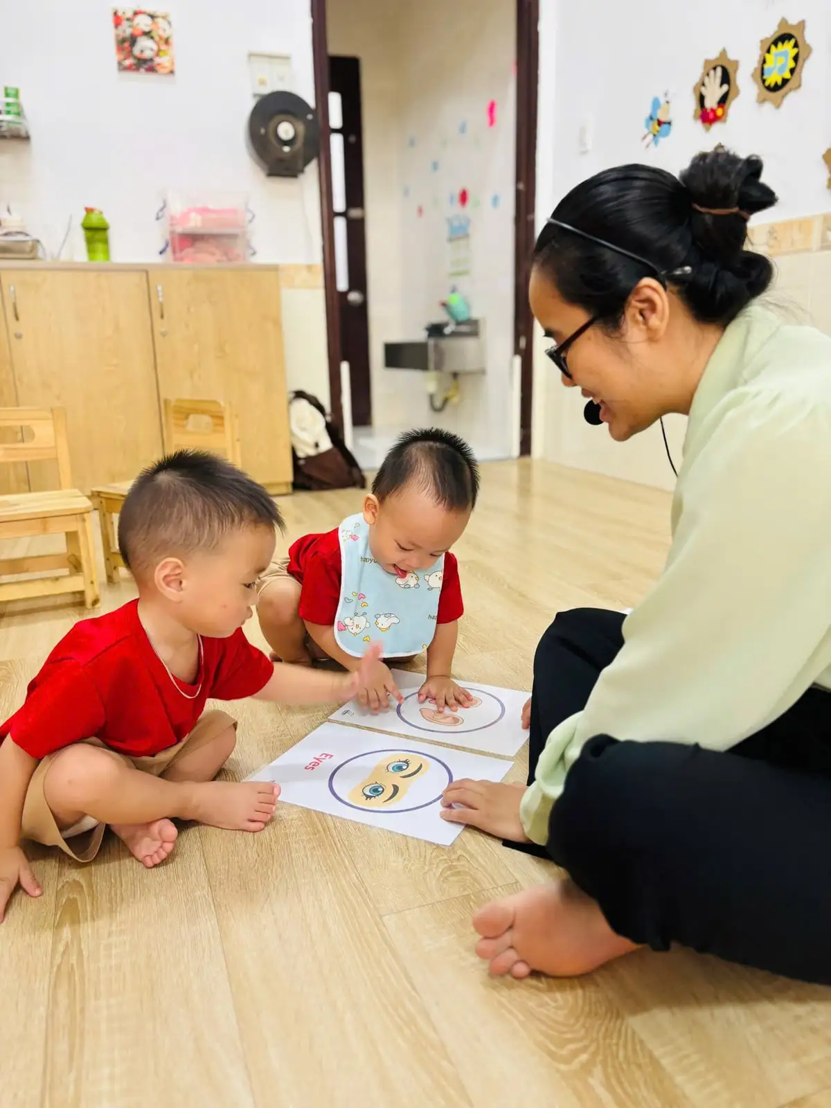 Cô giáo cười cùng hai bé lớp PreK1 chạm thẻ hình đôi mắt ở nhà trẻ Quận 7 JumboKiz