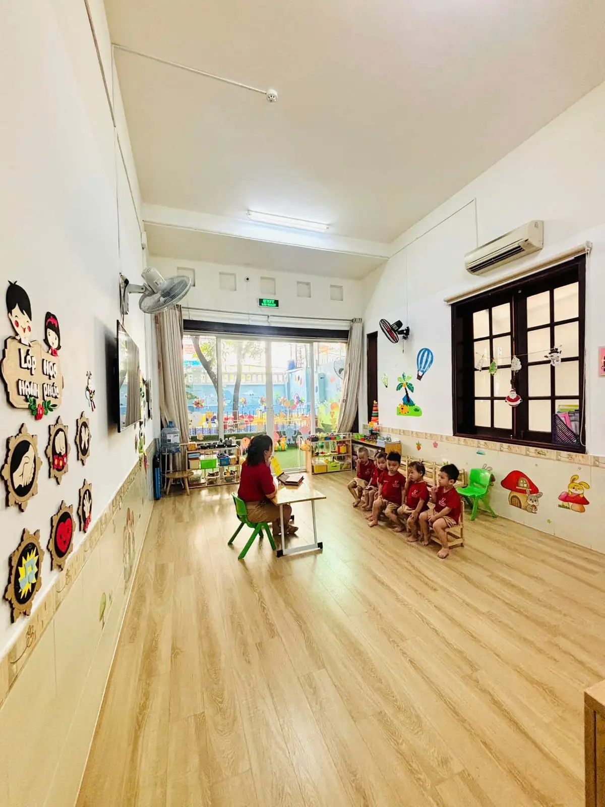 Toàn cảnh phòng học sàn gỗ sáng, cô ngồi bàn trước các bé ngồi ghế