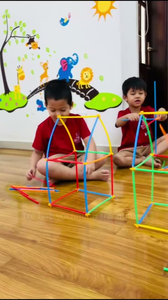 Hai bé trai mặc đồng phục đỏ ngồi trên sàn gỗ lắp khung bằng que nhựa nhiều màu, ảnh bìa video một ngày đi học ở trường mầm non quận 7 JumboKiz